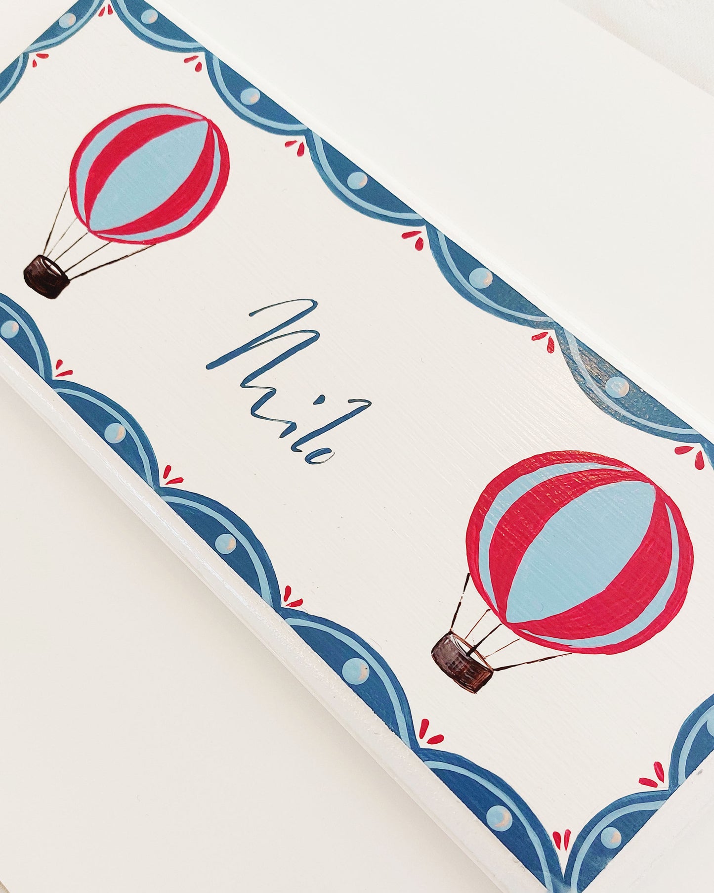 Nursery door sign personalised with the name 'Milo', featuring red and blue hot air balloons and a decorative blue scalloped border, perfect for baby room decor.