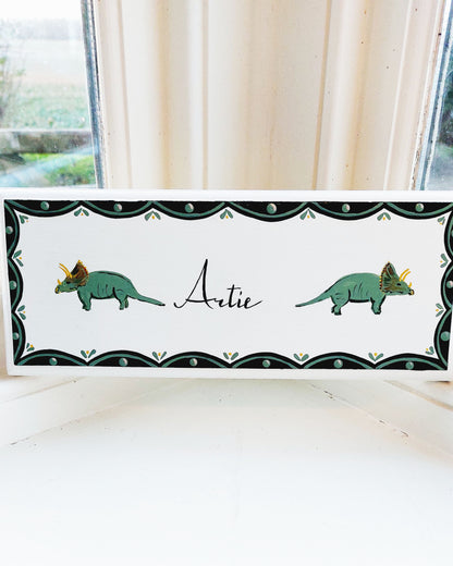 Nursery door sign personalised with the name 'Artie', featuring historic dinosaurs and a decorative blue and green scallop border, perfect for baby room decor.