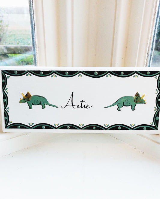 Nursery door sign personalised with the name 'Artie', featuring historic dinosaurs and a decorative blue and green scallop border, perfect for baby room decor.