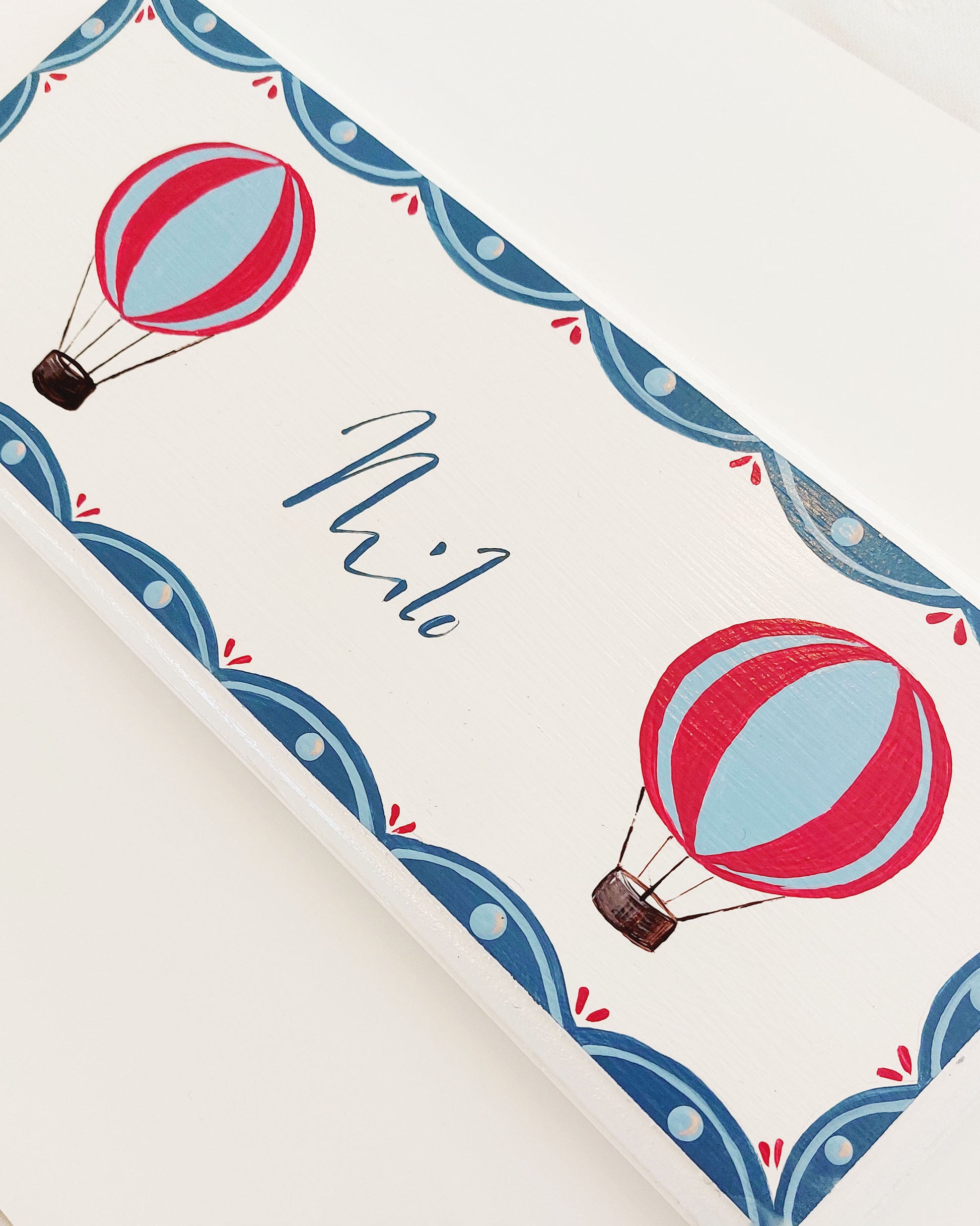 Nursery door sign personalised with the name 'Milo', featuring red and blue hot air balloons and a decorative blue scalloped border, perfect for baby room decor.